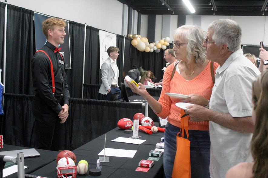 Luverne High School junior Jaydon Johnson (far left) talks with customers about his “Grand Prairie Sports” sports trivia cards Wednesday, May 7, at the 2025 CEO Trade Show as Kathy and Ron Vander Lugt of Chandler examine the cards created for Southwest Minnesota Christian High School in Edgerton. Kathy said she holds the school’s high jump record and wondered if her accomplishment was one of the trivia questions. The couple bought the set of trivia cards. Mavis Fodness/ Rock County Star Herald Photo