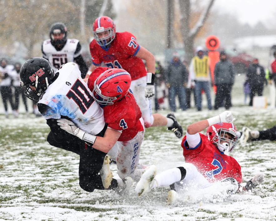 Samantha McGaffee photo: Patriot junior Beau Bakken (4) wraps up and tackles a Knights runner with sophomore Micha Bush (7) slipping on the cold, wet, snow-covered field and junior Jack Moser (71) closing in on the play. H-BC beat the Knights 28-0 at home Saturday, Oct. 28.