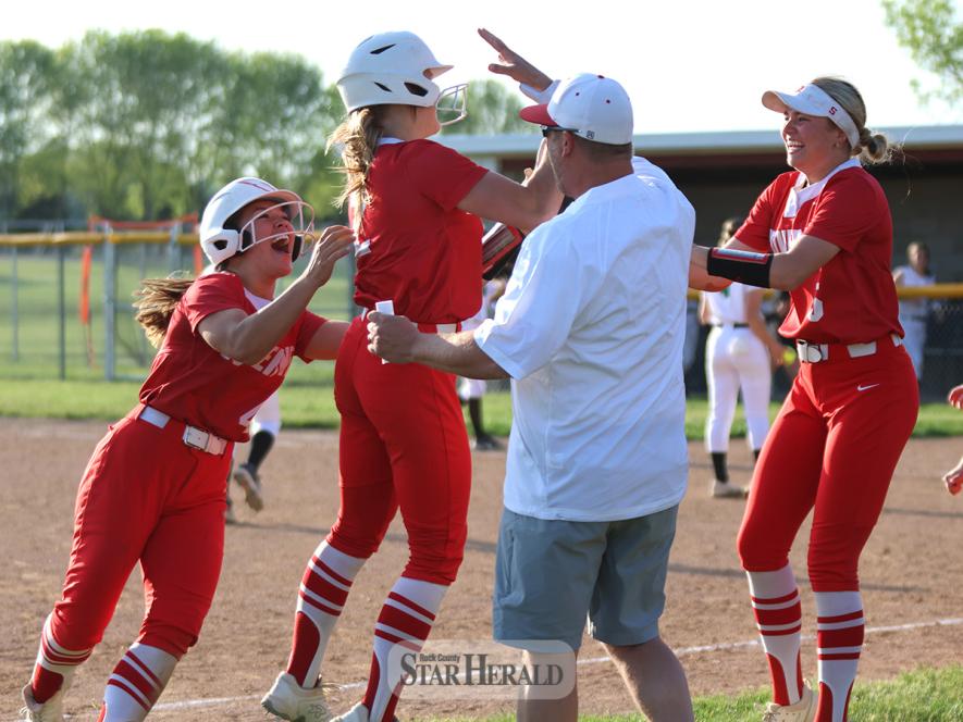 Senior Belle Smidt (left), junior Ella Reisdorfer (middle), coach Brooks Maurer and senior Tori Serie (right) celebrate Reisdorfer’s triple that sent Smidt across home plate for the 7-6 win over Pipestone Thursday, May 16, in Luverne.